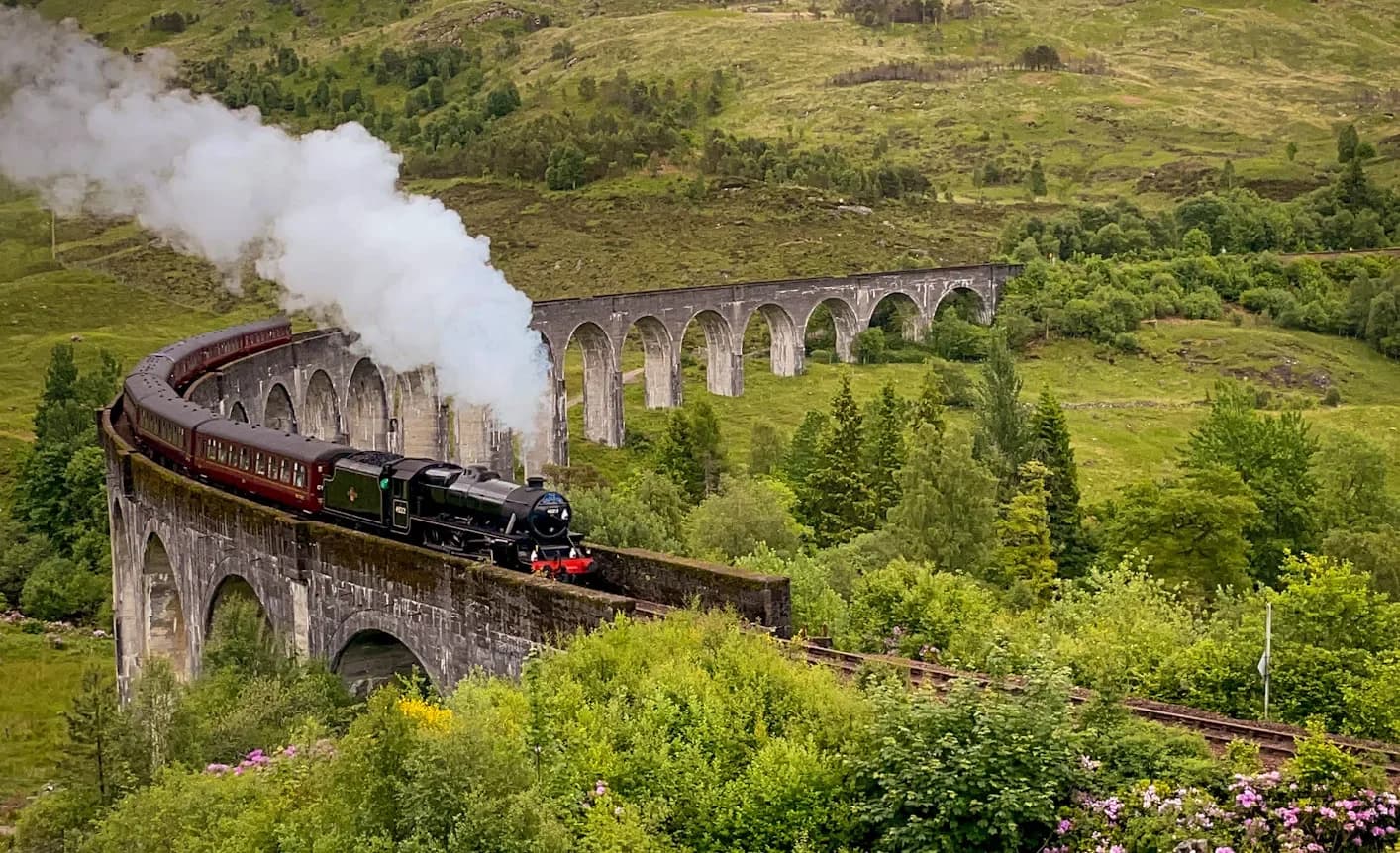 A steam train crossing the curved Glenfinnan Viaduct amid green Highland slopes