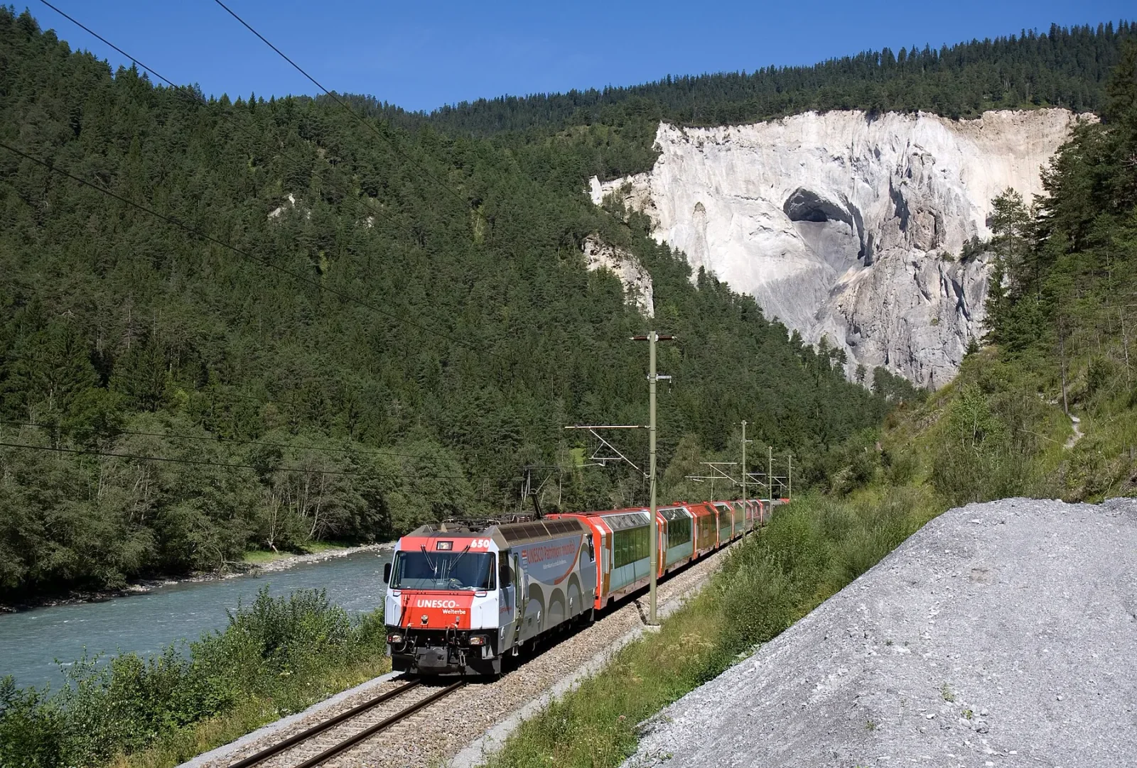 Two red Glacier Express trainsets following the Rhine through a pale-walled gorge